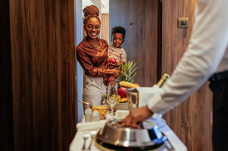 Wealthy hotel guest receiving room service. An African American hotel guest is with her son at the apartment door, she is being catered with lavish room service by the waiter.