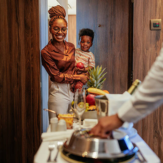 An African American hotel guest is with her son at the apartment door, she is being catered with lavish room service by the waiter.